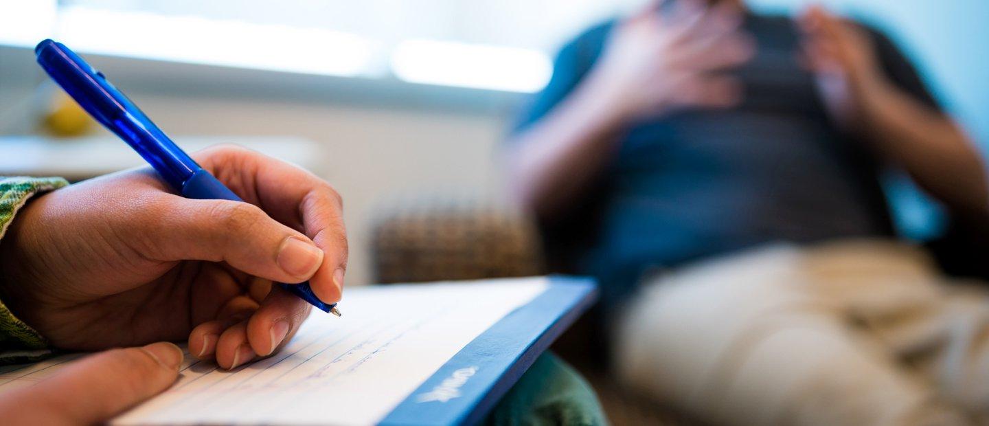 A hand holding a blue pen, writing on a notepad. Another person gesturing with his hands in the background.